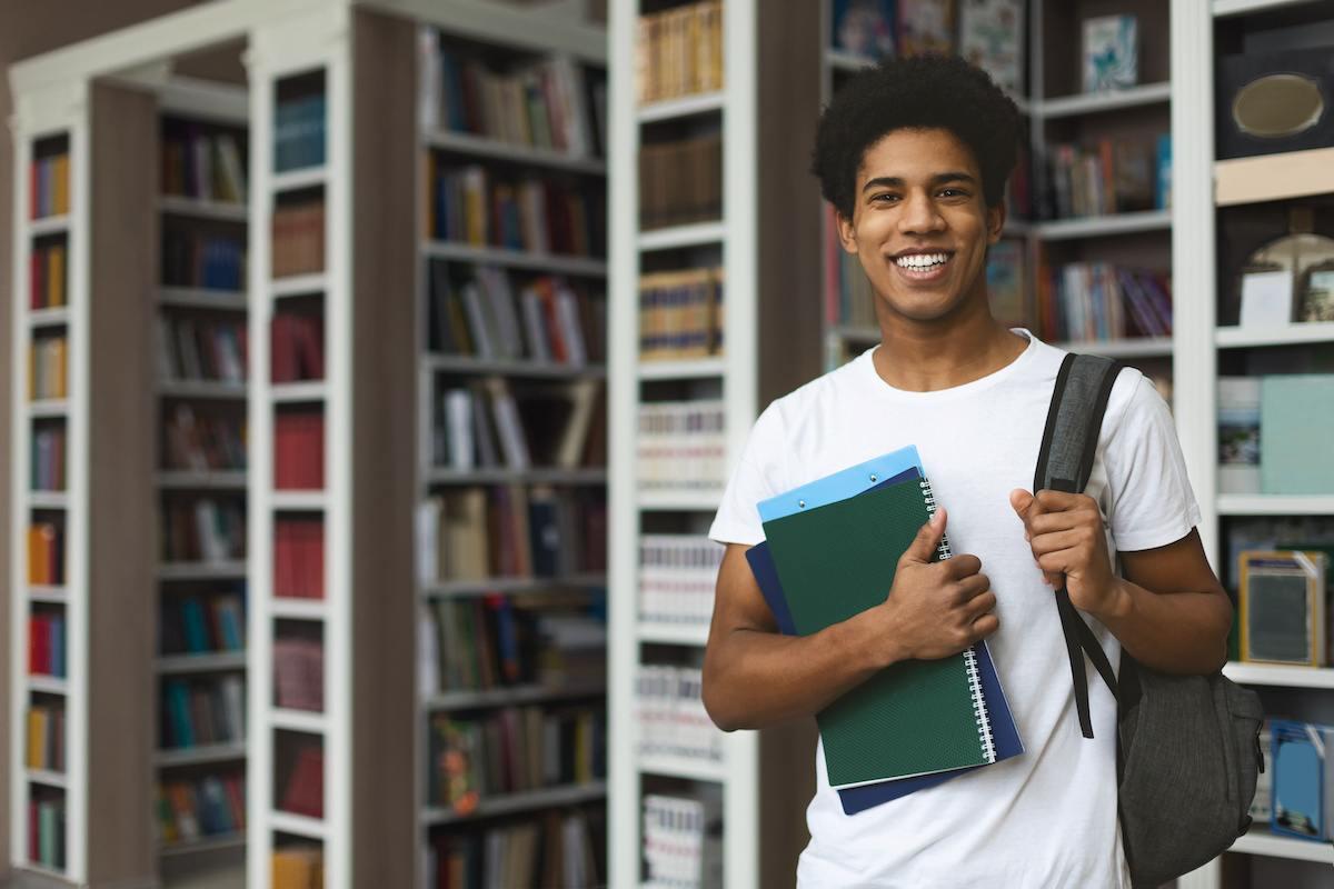A future North Carolina teacher standing in his campus library. He’s wearing a white T-shirt and holding books in one hand and a backpack on his shoulder with the other. 